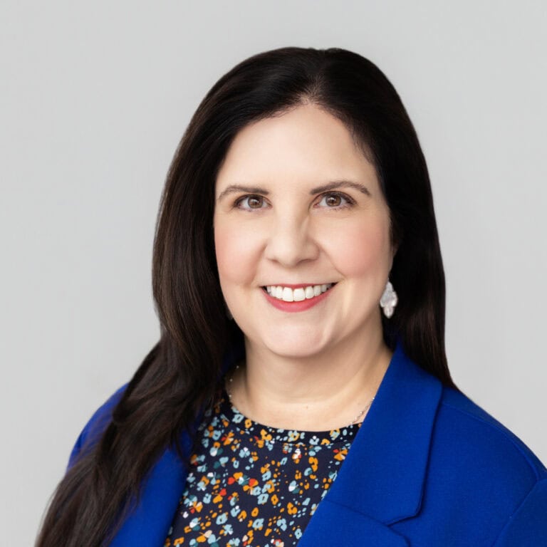 Smiling woman with long dark hair wearing a blue blazer and floral top against a light gray backgrou.