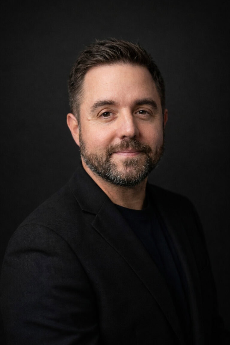 A man with dark hair and a beard, wearing a black blazer, smiling softly against a dark background.