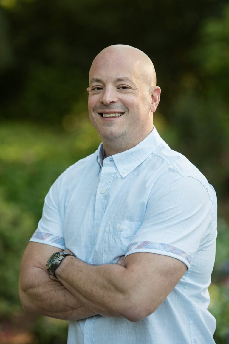 Smiling bald man in light blue shirt standing outdoors with arms crossed.