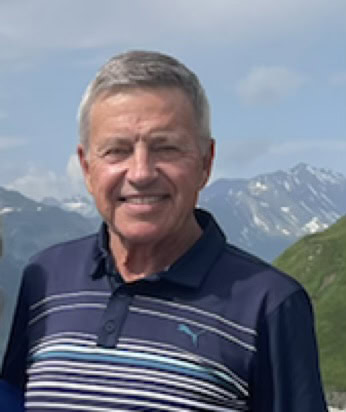 A man with gray hair smiling outdoors with mountains in the background.