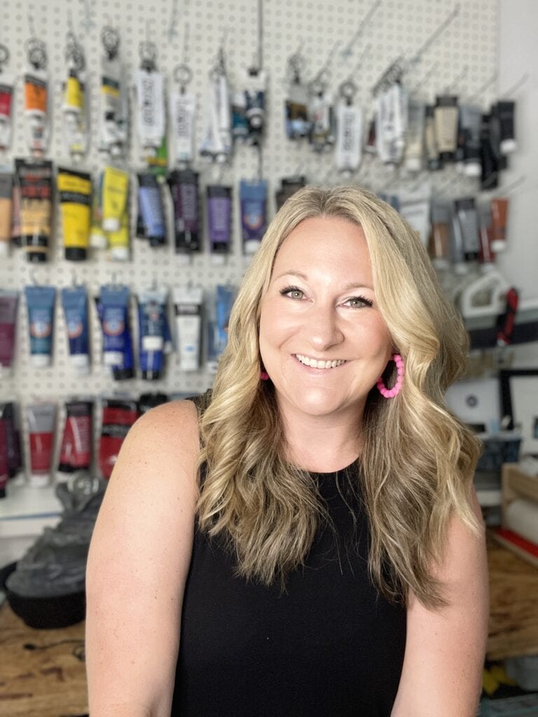 Woman with blonde hair and pink earrings smiling in art supplies store.