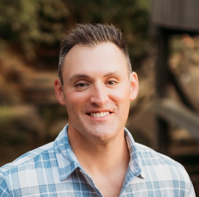 Smiling man in blue plaid shirt outdoors with blurred background.