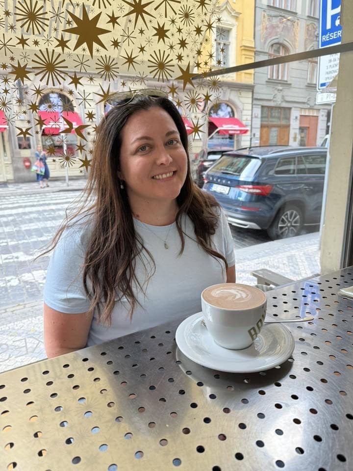 A woman sitting at a cafe table with a cup of coffee, smiling at the camera.