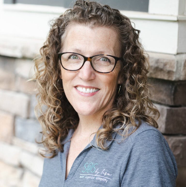 Woman with curly hair and glasses smiling outdoors in front of a stone wall.