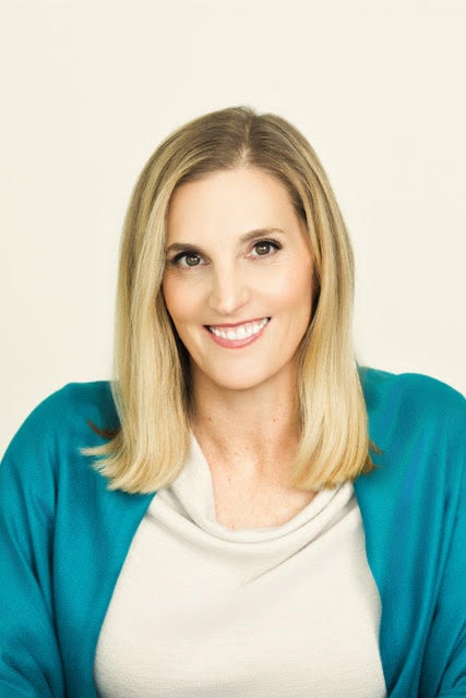 A woman with blonde hair smiling at the camera, wearing a white top and a teal cardigan.