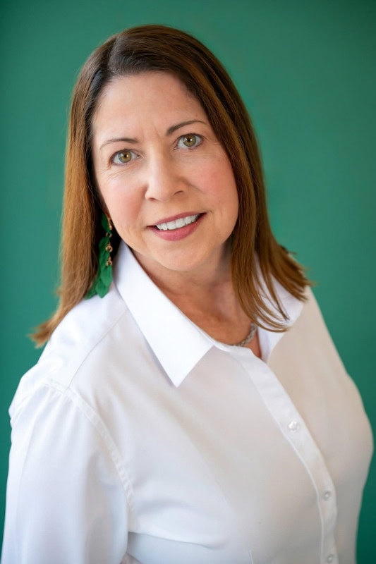 Woman with brown hair wearing a white shirt and green earrings against a green background.