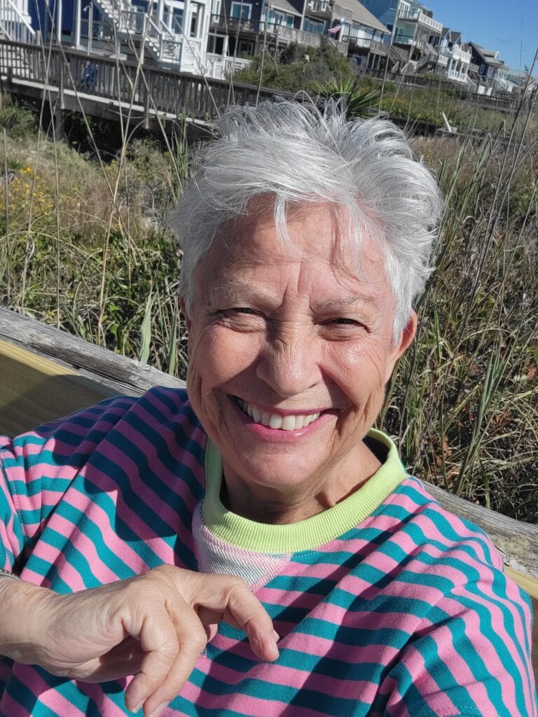 A woman with short gray hair smiling outdoors near a wooden walkway and modern houses.
