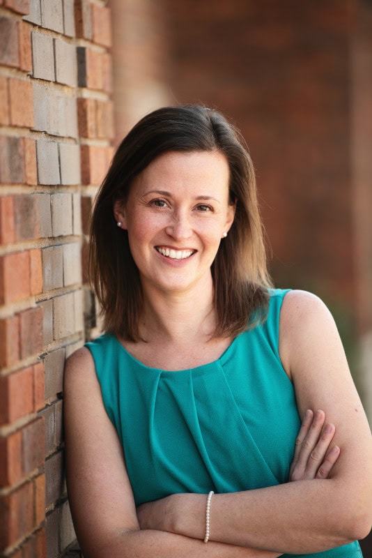 Smiling woman with shoulder-length brown hair in a teal sleeveless top leaning against a brick wall.