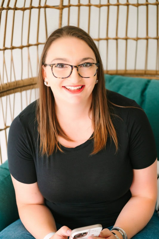 Woman with glasses smiling, holding a smartphone, sitting on a green cushion.