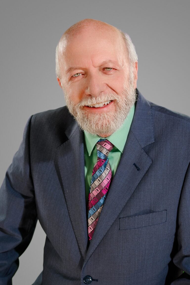 A smiling older man with a beard wearing a dark suit, green shirt, and colorful tie against a gray b.